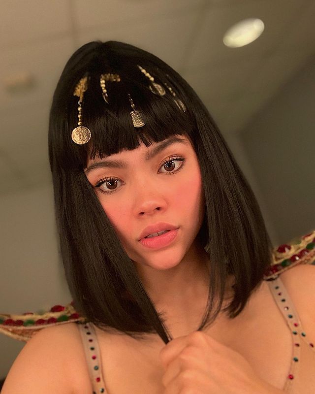 Myra Molloy in a fashionable golden jewelry and thin strapped light-brown dress.